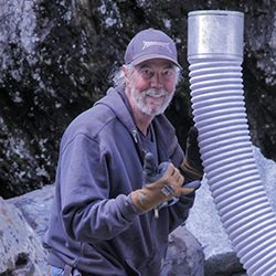 Um homem com barba cinza e um boné de beisebol em um ambiente industrial.