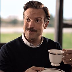 Homem de bigode segura uma xícara de chá e sorri.