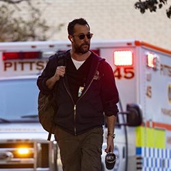 Homem com óculos escuros e mochila caminhando em frente a uma ambulância.