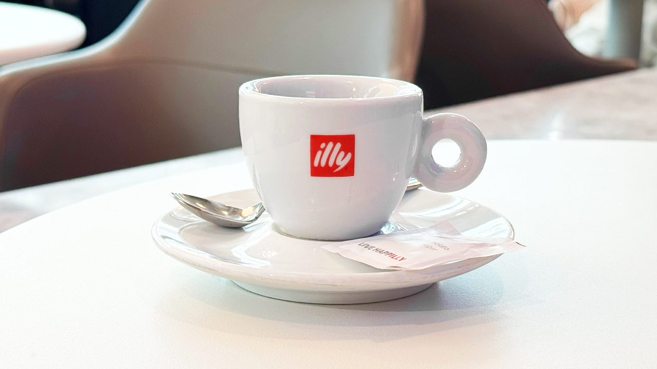A white illy coffee cup with a red logo sits on a saucer with a spoon and sugar packet on a café table.
