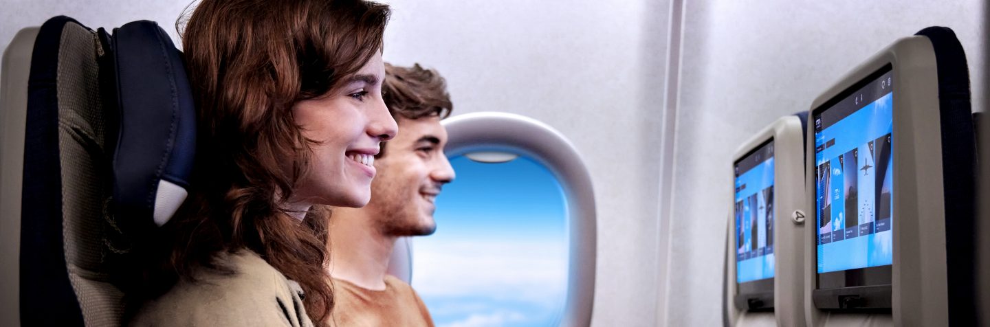 Un homme et une femme assis côte à côte dans un avion, souriant en regardant la télévision à bord, installée sur les sièges devant eux.