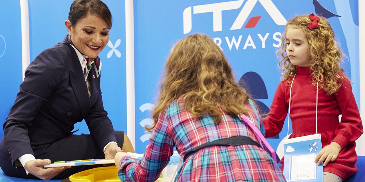An ITA Airways staff member assists two children in a dedicated airport area.