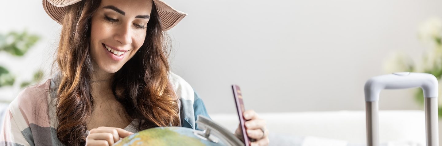 Donna sorridente con un cappello tiene un passaporto e indica un mappamondo.