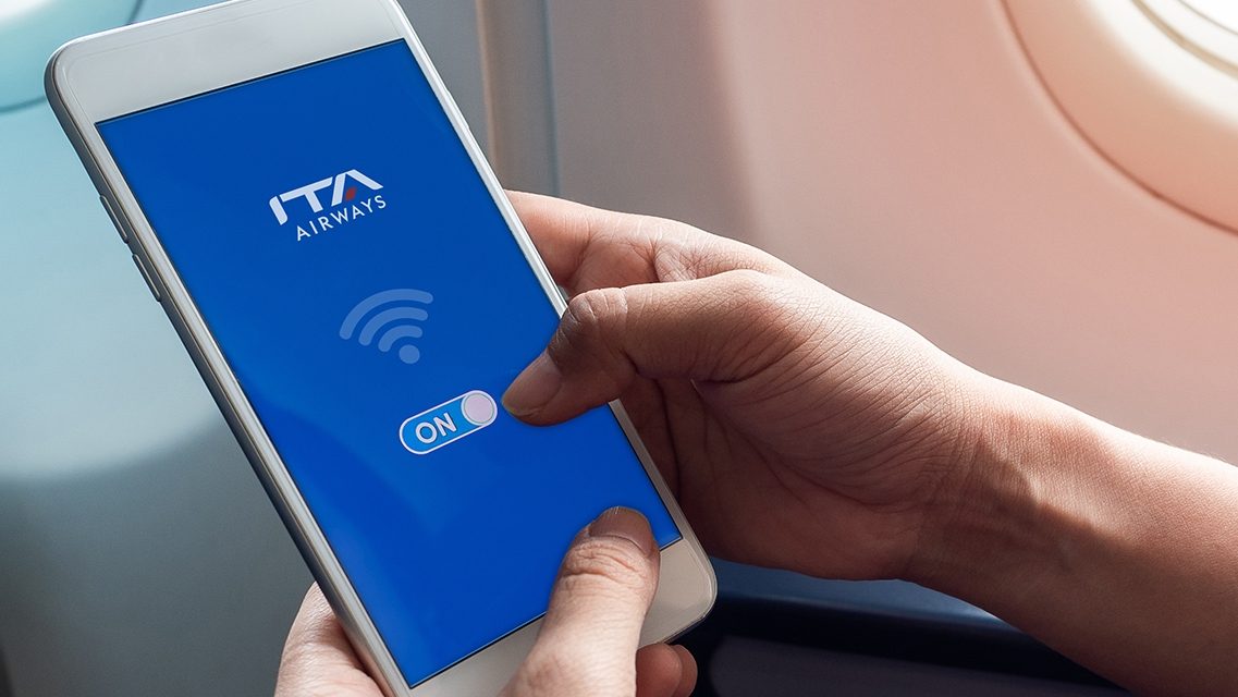 A hand holding a smartphone showing the ITA Airways logo and a Wi-Fi symbol on board the aircraft.