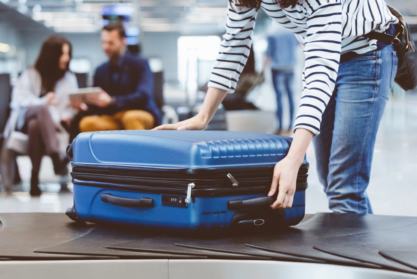 Young woman passenger collecting her luggage from conveyor belt. Female traveler picking up suitcase from baggage claim line in airport terminal.