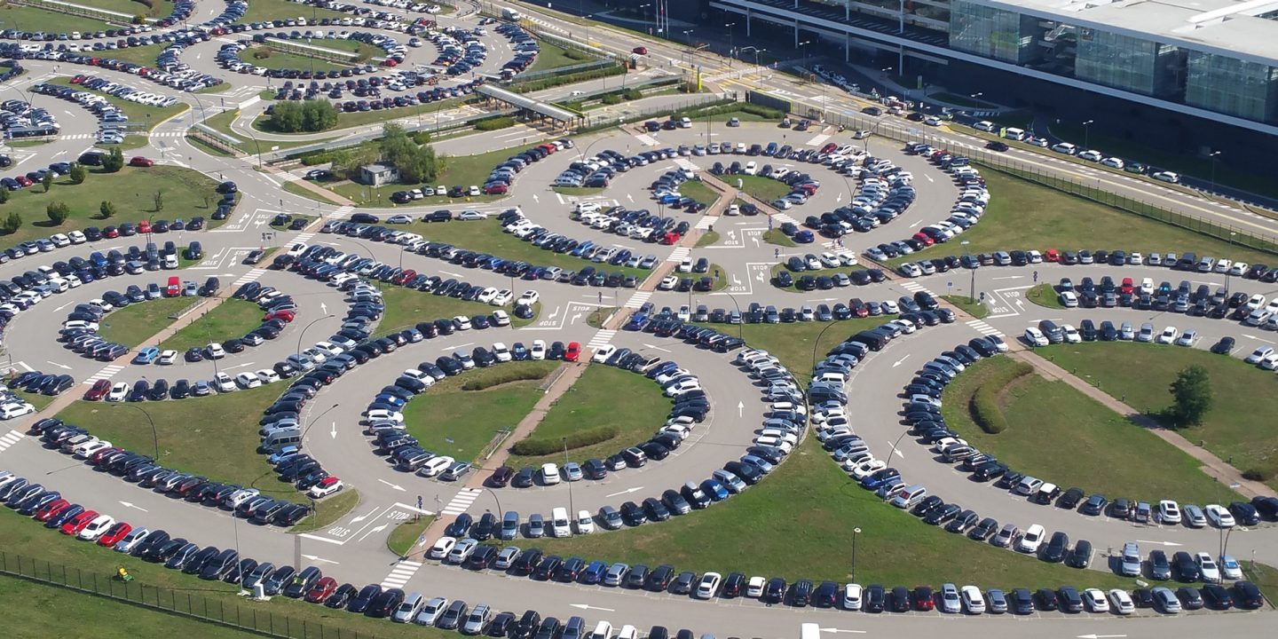 Vue aérienne d’un parking aéroportuaire avec des voitures disposées en sections circulaires près du terminal.