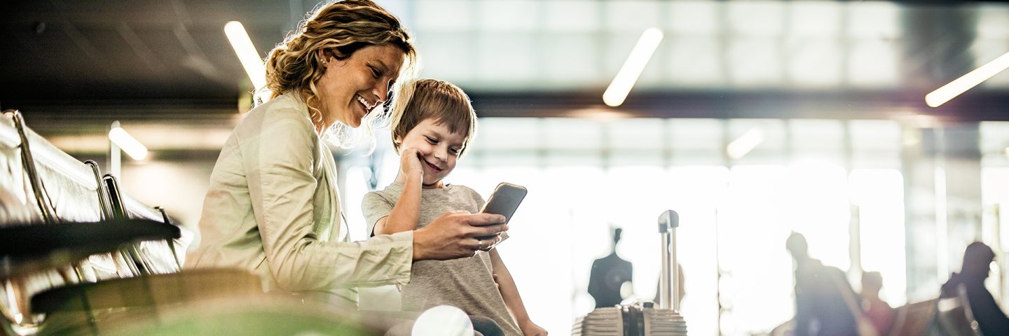 Une mère et son fils rient ensemble en regardant un téléphone, assis dans la zone d’attente de l’aéroport.