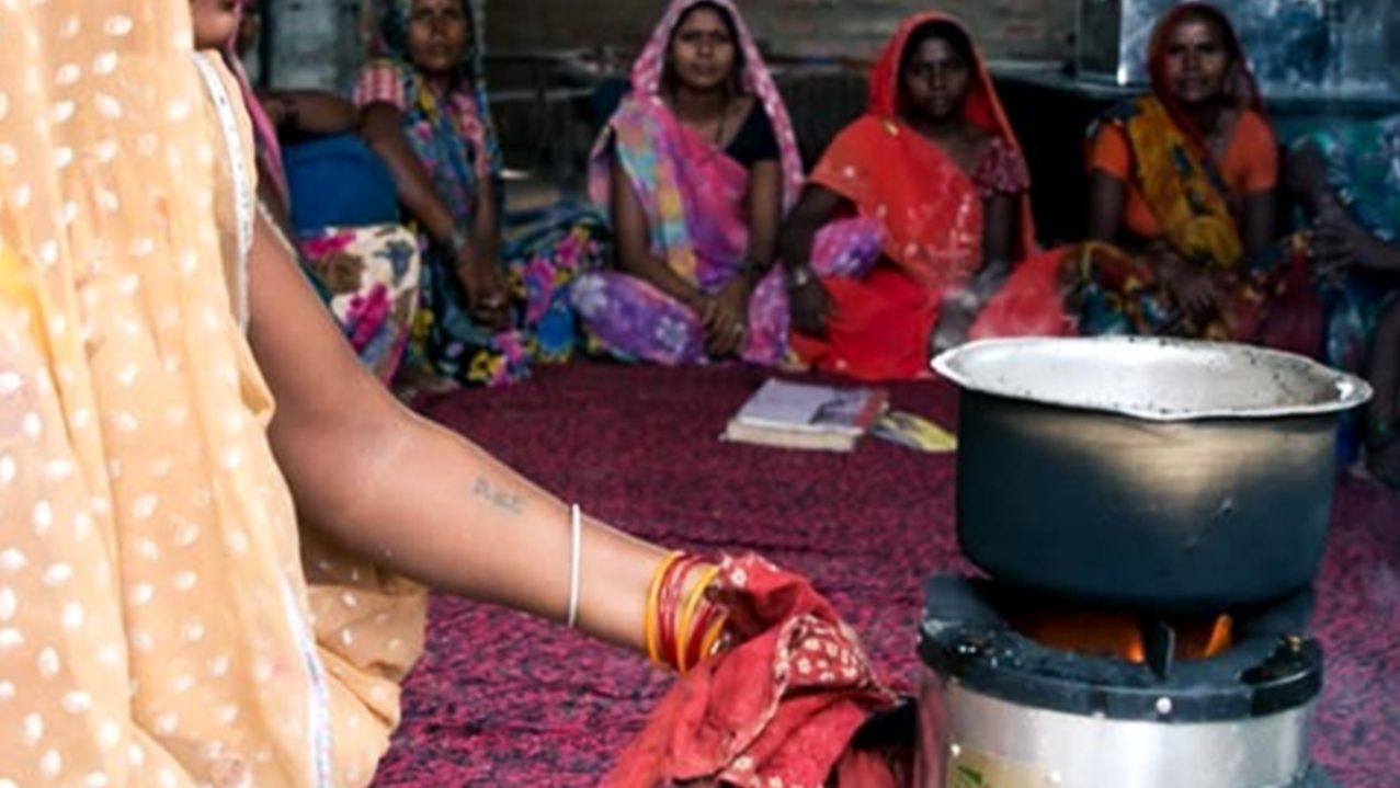 Una donna in sari prepara un piatto mentre un gruppo di donne la guarda con interesse.