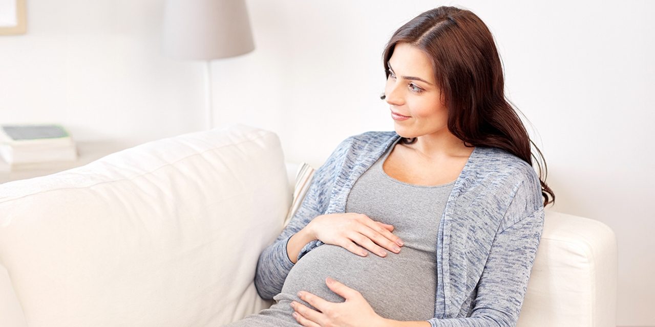 A pregnant woman sitting on a sofa whilst holding her pregnancy bump.
