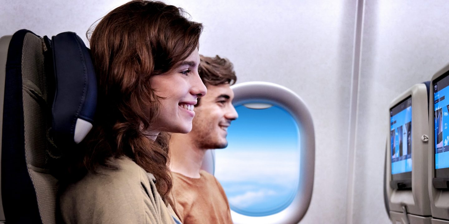 A man and woman sitting side-by-side on a plane smile while watching in-flight TV on the screens in front of them.