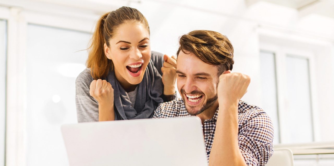 Two people sitting at a table with a laptop, both raising fists in a celebratory gesture.
