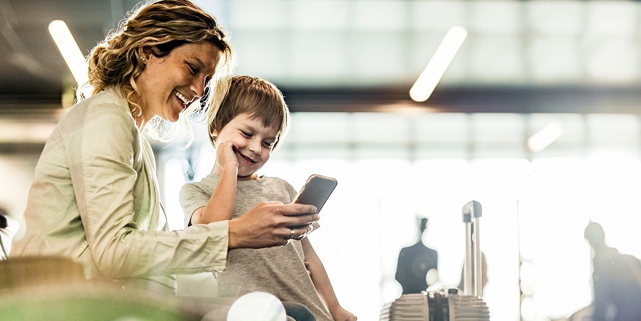A mother and son laughing together whilst looking at a phone and sitting in the airport waiting area.