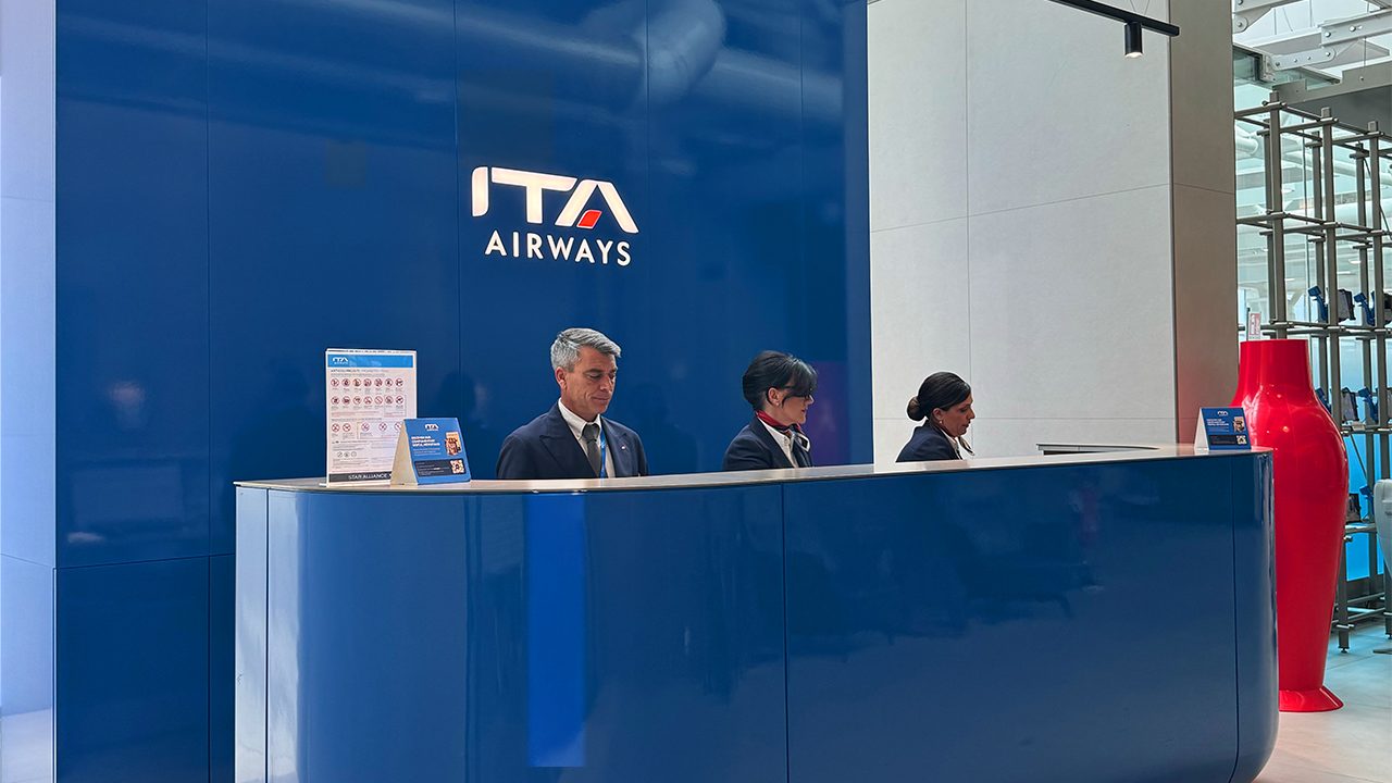 Three airline staff in formal attire stand behind a sleek blue ITA Airways counter.