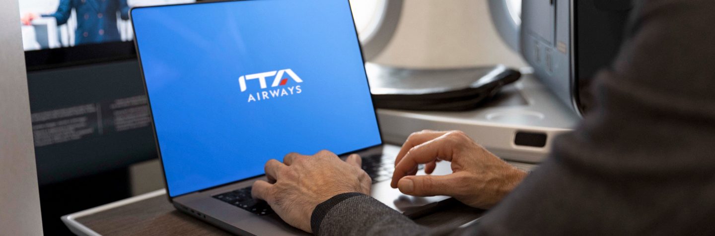 Close-up van iemand die op een laptop typt, met op het scherm het ITA Airways-logo tegen een blauwe achtergrond.