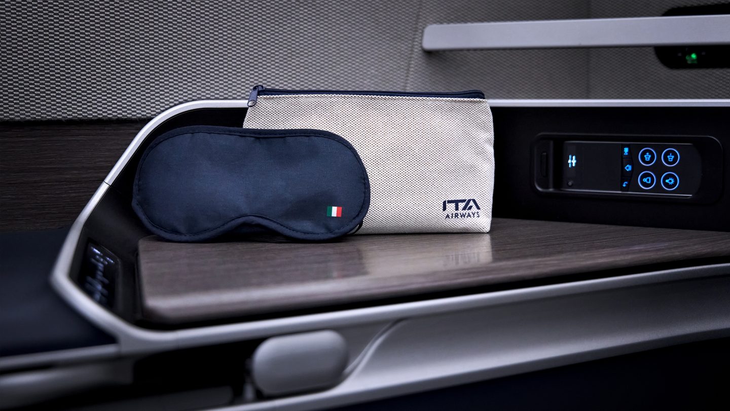 Máscara de dormir ITA com a bandeira italiana bordada e nécessaire com o logotipo da ITA dispostas sobre a mesa.